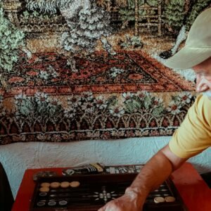 Elderly man enjoying backgammon beside a colorful tapestry backdrop.