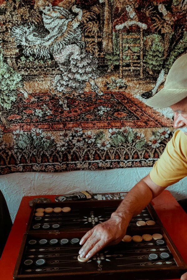 Elderly man enjoying backgammon beside a colorful tapestry backdrop.
